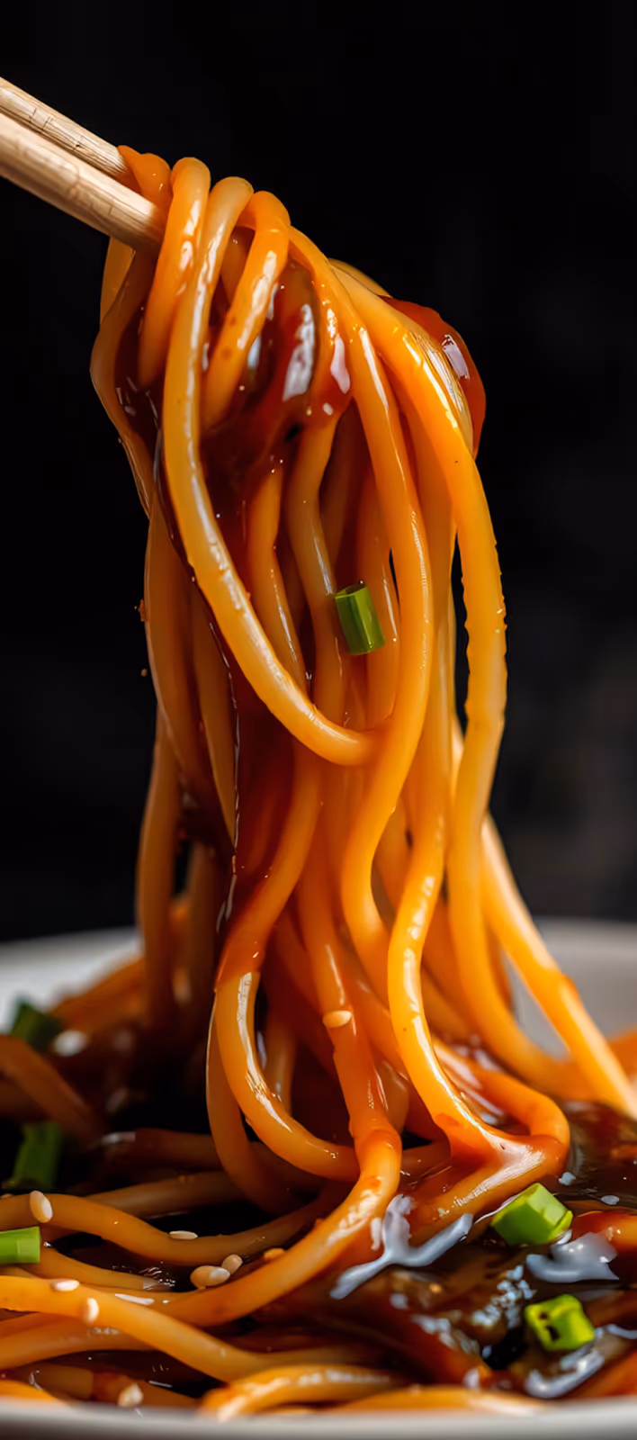Chopsticks lifting spaghetti noodles coated in a glossy red sauce with sesame seeds and green onion garnish.